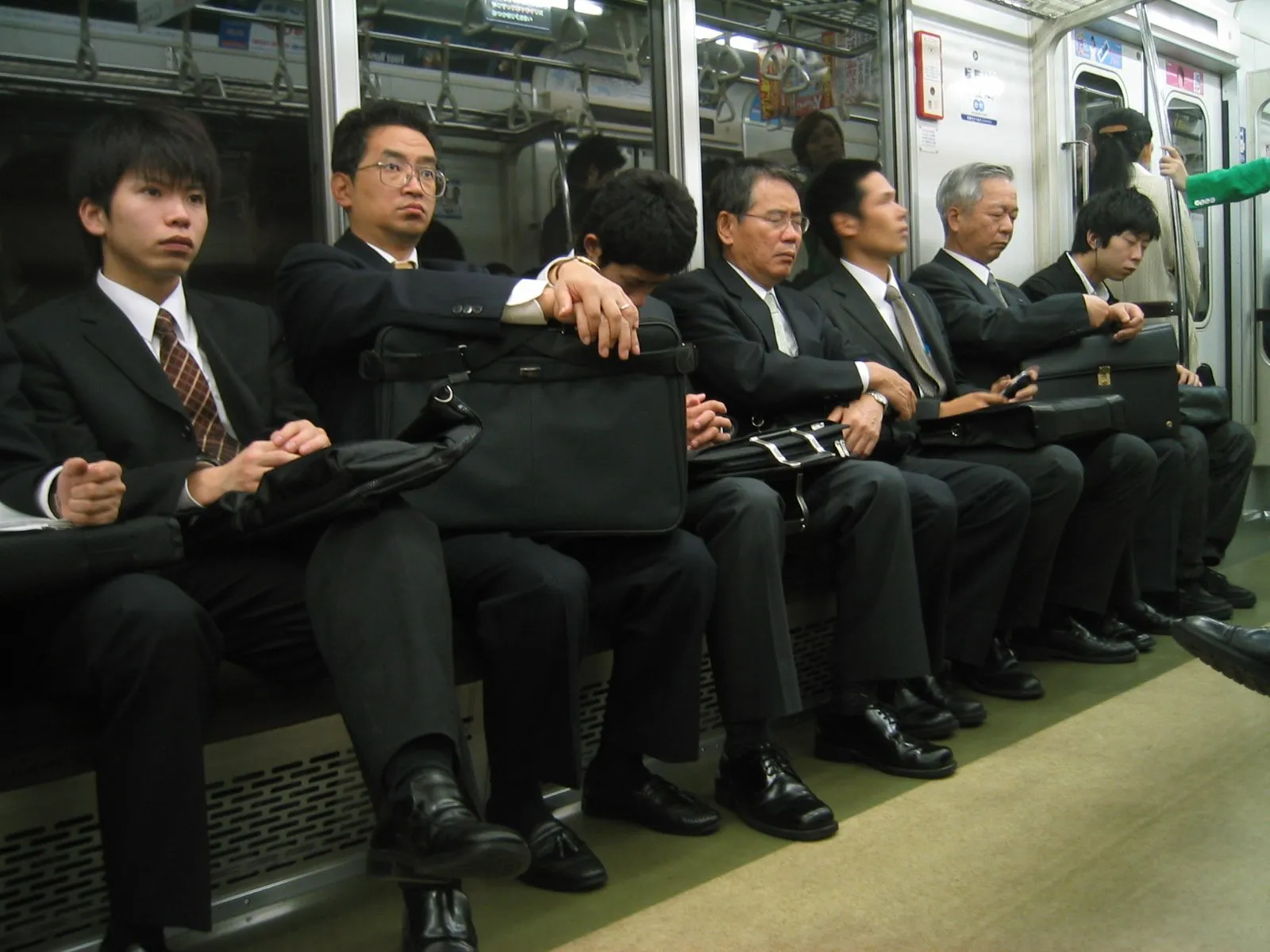 Hombres de negocios con traje oscuro sentados en un tren, algunos con bolsos de mano, otros usando teléfonos móviles, todos con expresión seria y durmiendo, en vagón del metro.