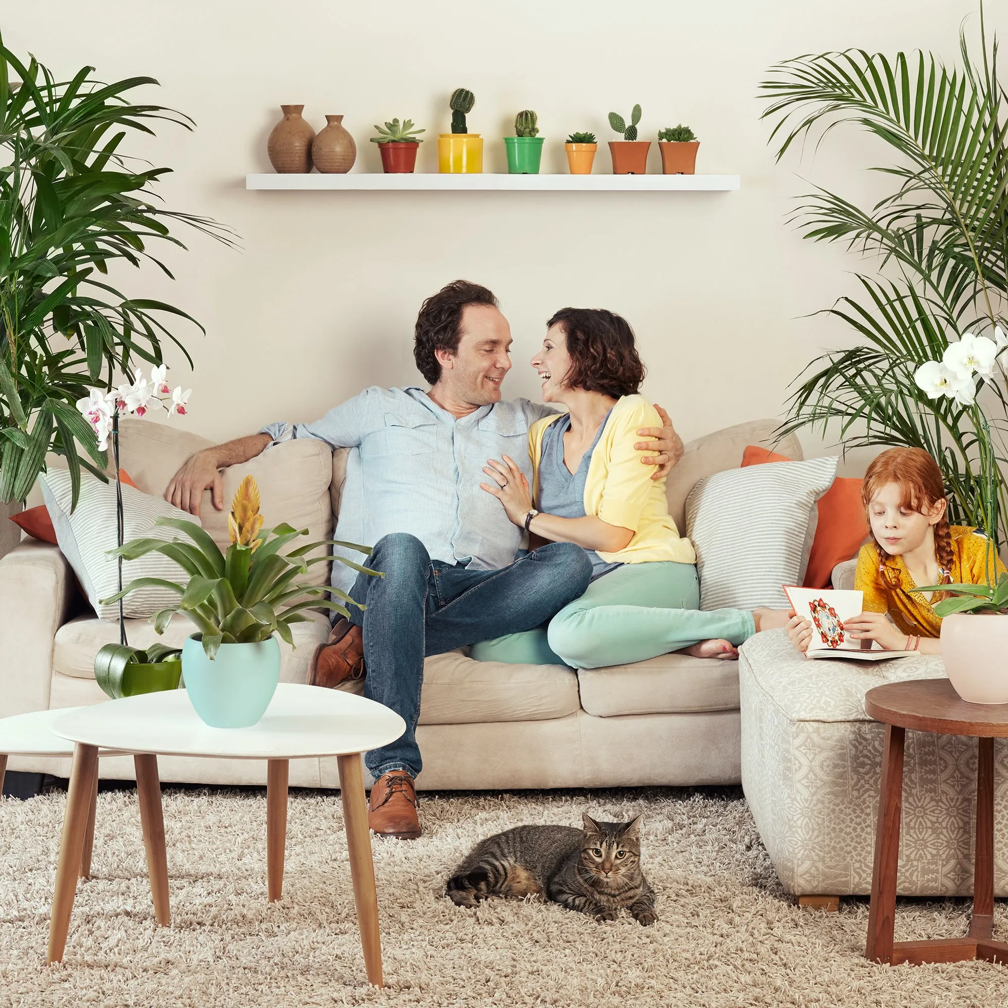 Una familia feliz en la sala de estar rodeada de plantas y decoraciones, con un gato tumbado en la alfombra y una niña leyendo un libro.