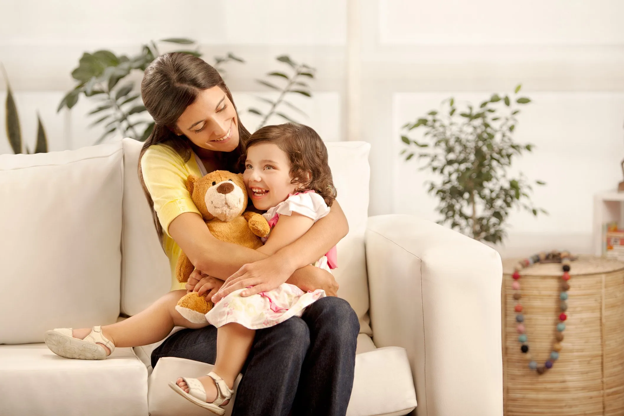 Una madre y su hija pequeña abrazándose y sonriendo en un sofá, con la niña sosteniendo un oso de peluche, en un ambiente hogareño con plantas y decoración