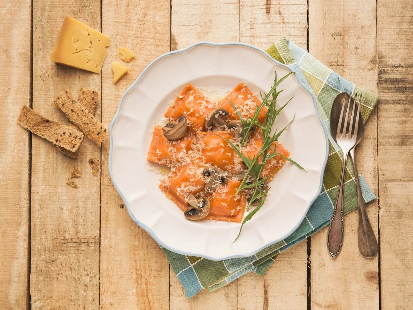 Plato de pasta con salsa de tomate, queso rallado, champiñones, y rúcula en un plato blanco sobre una mesa de madera con tenedor y cuchara en un paño de tela. A la izquierda, queso, pan integral y galletas. Consiste en la comida italiana.