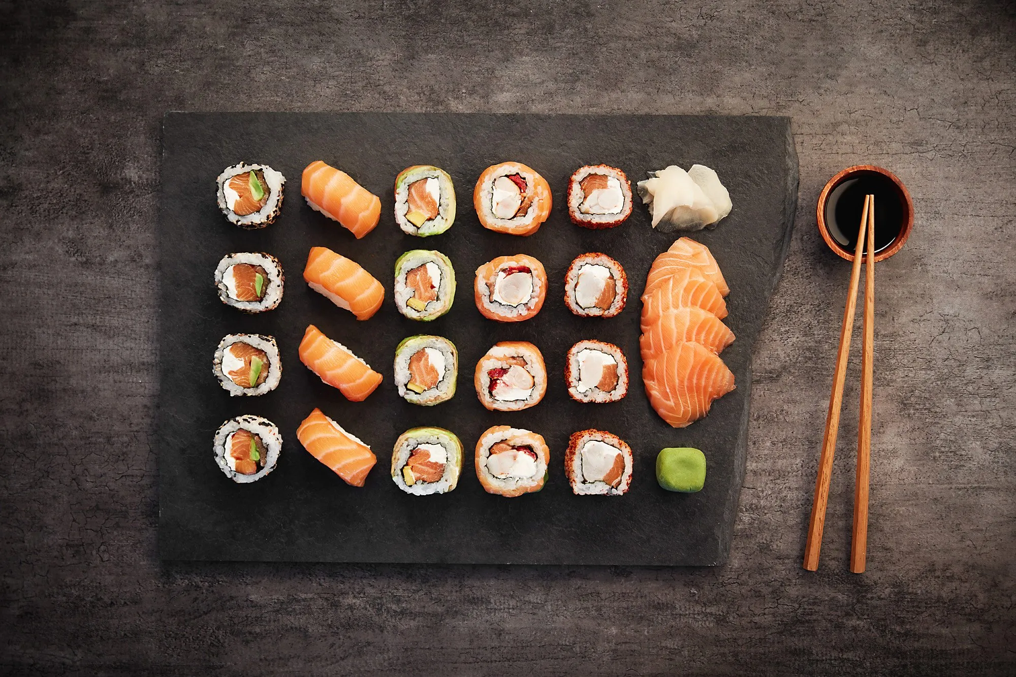 Plato con variedad de sushi en una bandeja negra, acompañado de jengibre, wasabi, y salsa de soja en un tazón con palillos chinos.