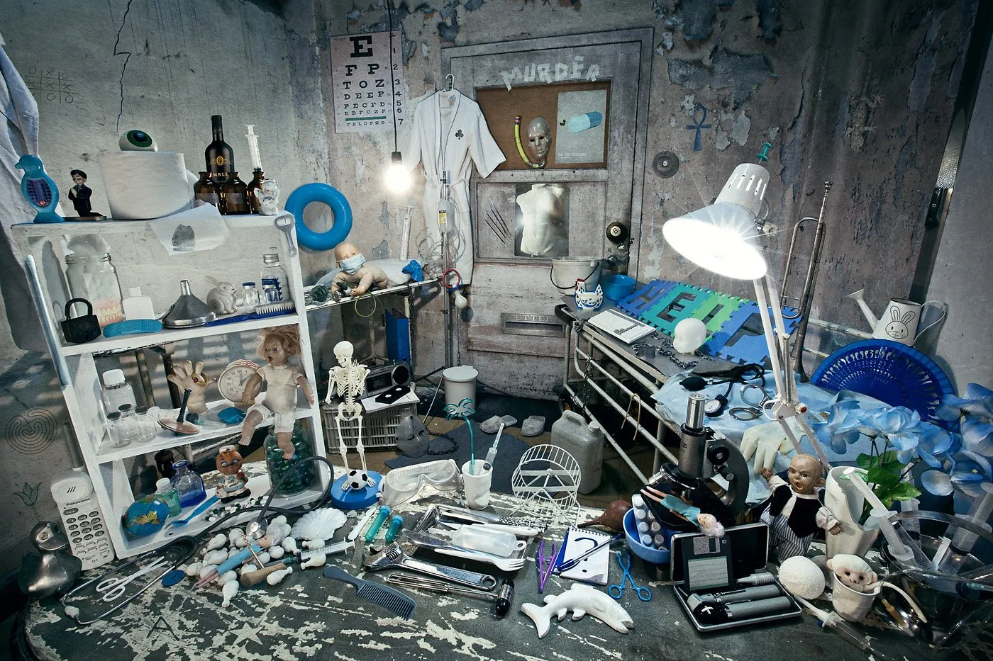Un espacio caótico lleno de objetos diversos como muñecos, instrumentos médicos, libros, una lámpara, y objetos de oficina, en un entorno que parece un cuarto desordenado o laboratorio improvisado.